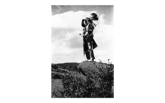 Person stands on a rock wearing a feathered headdress, looking into the distance. Vast sky and hills form the background, evoking a serene mood.