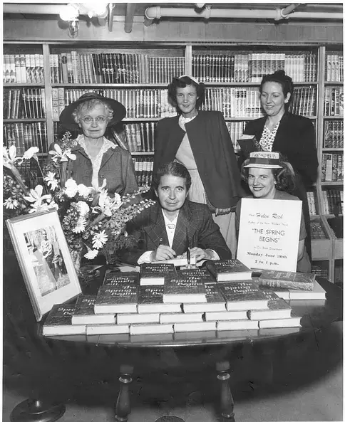 Woman signing books at a table with flowers; four women stand behind her in a library. A sign promotes "The Spring Begins" event.