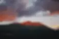 Mountain peak bathed in orange sunset light under dramatic clouds. Dark foreground contrasts with vibrant sky, creating a serene mood.