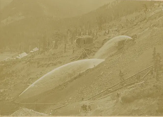 Hillside with cascading water streams for hydraulic mining; rustic cabins and dense forest in the background; sepia tones create a historical feel.