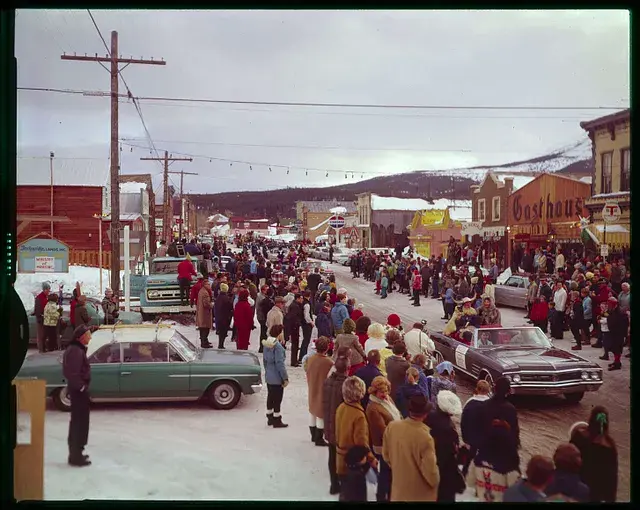 A winter street parade with crowds bundled in coats; classic cars and floats pass by; signs for "Gasthaus" visible; snow-covered landscape.