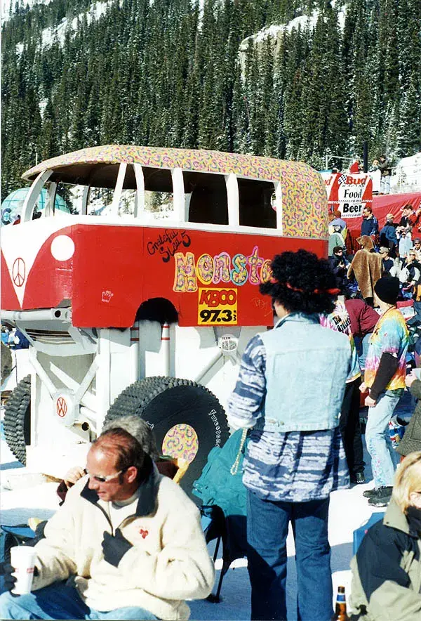 Red bus float with large wheels and colorful patterns, surrounded by a crowd in winter clothing. Mountains and trees in background. Text: "Monster KBCO 97.3".