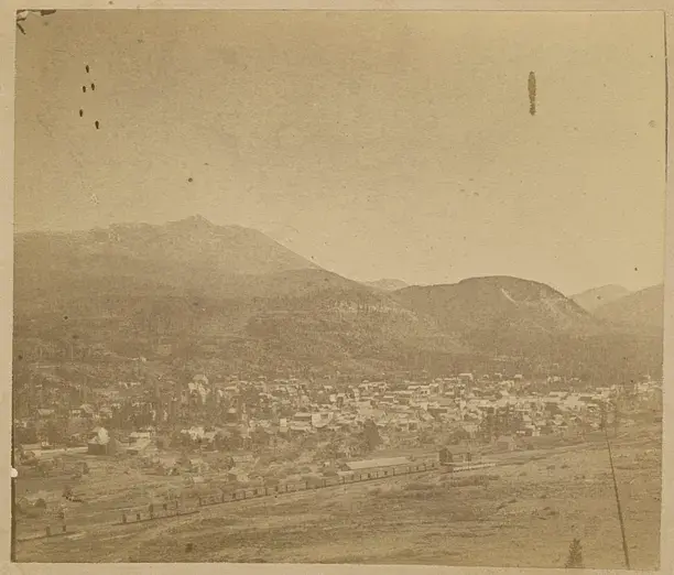 A panoramic view of a small town with clustered buildings, surrounded by mountains. A train runs along the foreground. Sepia-toned, peaceful scene.