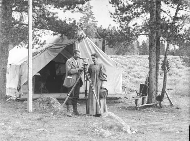Two people stand outside a canvas tent in a forest, using a theodolite. Trees surround them, creating a rustic, exploratory mood.