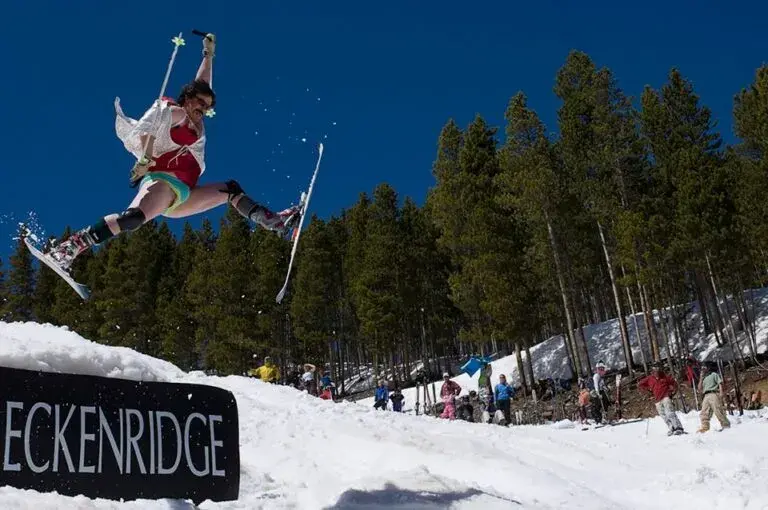 Skier in colorful attire jumps mid-air in snowy forest setting. "Breckenridge" sign visible. Energetic mood, clear blue sky.