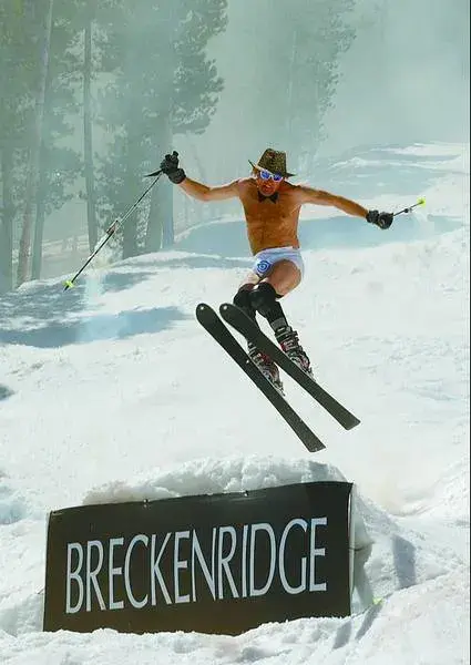 Skier in shorts and cowboy hat jumps over a snowy hill labeled Breckenridge, against a backdrop of trees, exuding fun and adventure.