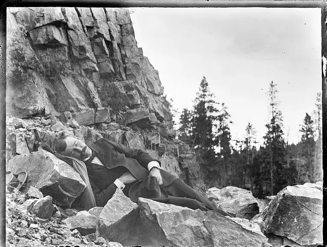Man in a suit sleeping on rocks near a cliff. Pine trees are in the background under a cloudy sky, conveying tranquility.