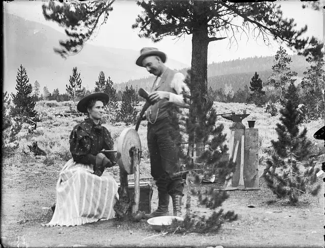 A woman uses a grinding wheel while a man in a hat looks on, in a forested rural setting. They appear focused in this vintage scene.