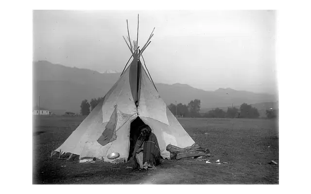 A person sits at the entrance of a tipi in an open field with mountains in the background, creating a serene and solitary mood.
