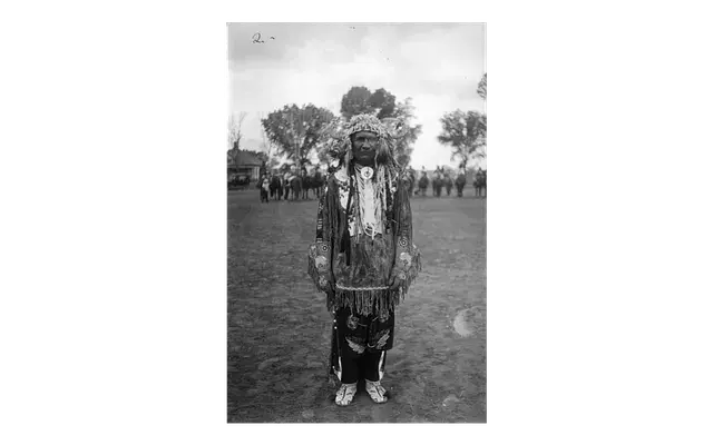 Man in traditional attire with headdress stands on grassy area. Trees and people in the background. Monochrome image conveys a historical mood.