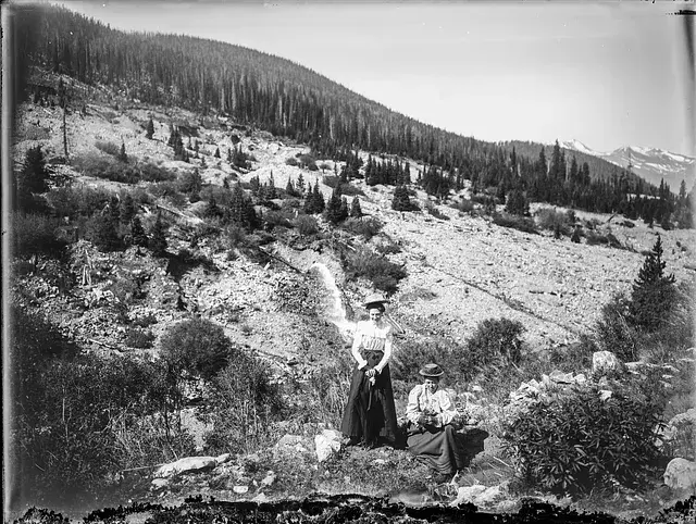Two people in vintage attire stand and sit in a mountainous landscape with scattered trees. One holds a hat. The scene is calm and serene.