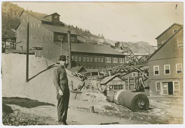 Man in dark clothing and hat stands in front of a large industrial building with a chimney. The setting is mountainous and rugged.