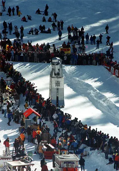 Crowd gathered on snowy hill watching luge run. People in colorful winter clothes. Focus on sled and track. Energetic winter scene.