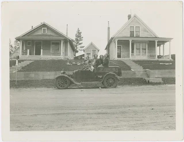 Vintage car with six people parked on a dirt road in front of two early 20th-century houses. The mood is relaxed and joyful.