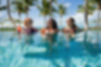 3 caucasian women in a pool holding drinks and laughing, palm trees in background.