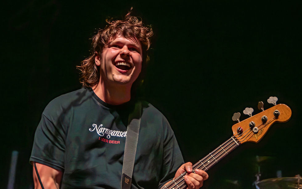 Smiling bassist on a dark stage wearing a Narragansett Lager Beer shirt, holding a bass guitar.