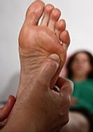 Reflexologist applying pressurer to the arch of a bare foot, with a client lying in the background.