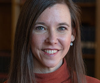 Image of a slim brunette woman with pale skin facing camera and smiling. She wears a rust colored turtleneck.