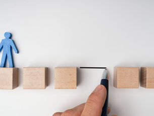 A blue wooden figure standing on blocks while a hand draws a bridge to the next step, symbolizing scaffolding bravery and supporting early career staff through the transition from school to work