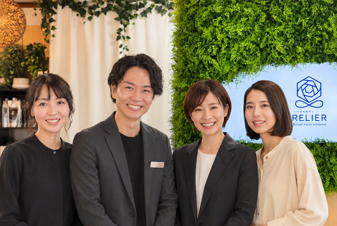 緑の壁と「RELIER」ロゴの前で、黒やベージュのスーツ姿の4人が笑顔で並ぶ、明るい店内の記念写真。
