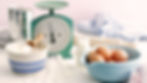 Vintage kitchen scene with a scale, flour bowl, eggs in a blue bowl, milk bottle, and rolling pin on a white cloth. Soft light, baking mood.
