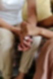 Two people seated, one in a white shirt and beige pants, holding hands in a comforting gesture. Background shows a striped cushion.