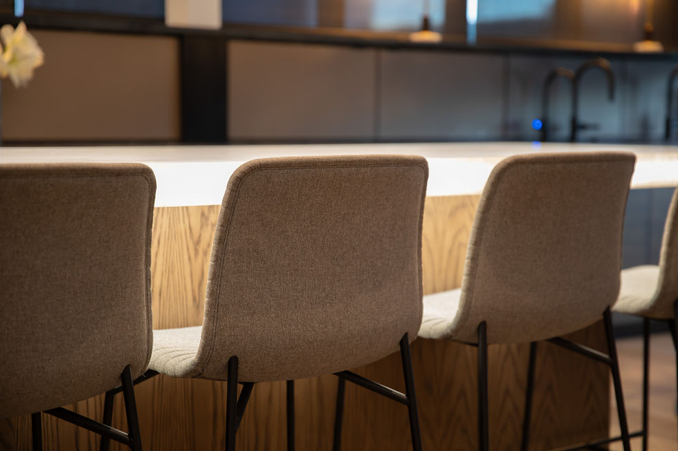 Bar with three beige upholstered chairs facing a backlit wooden counter in a modern kitchen. Dark cabinets and a faucet in the background.