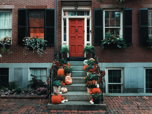 Red brick house with a bright red door, flanked by windows with black shutters. Steps adorned with pumpkins and flowers, creating a cozy vibe.
