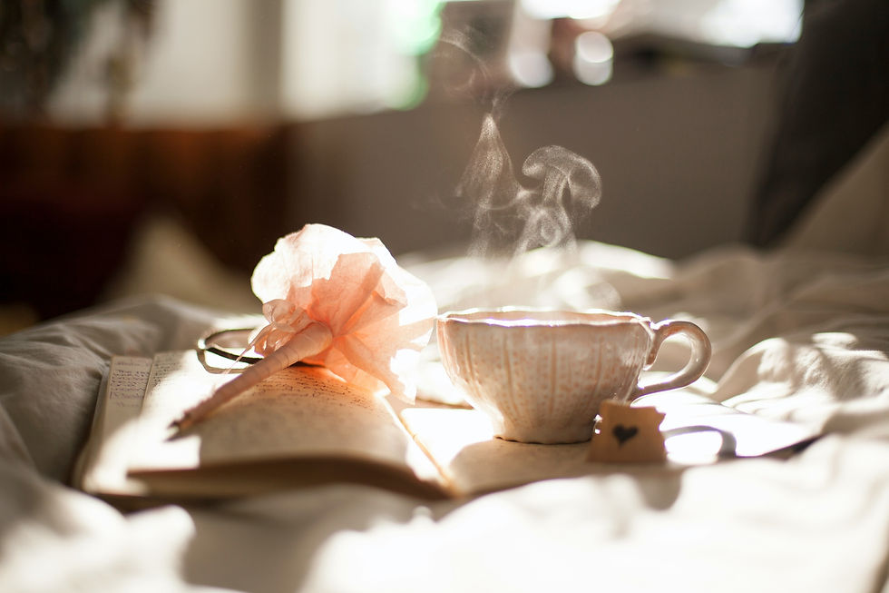 Steaming teacup, a journal, and a pink flower pen on a bed. Sunlight casts a warm glow, creating a cozy and serene atmosphere.