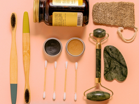 Toothbrushes, jade roller, gua sha, jar, creams, and cotton swabs on peach background. Natural skincare items neatly arranged.