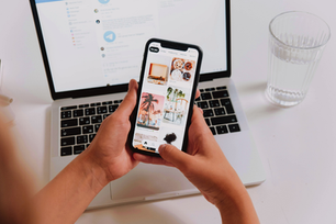 Hands holding a smartphone displaying photos, in front of a laptop with a messaging app. A glass of water is nearby. Cozy, work setting.