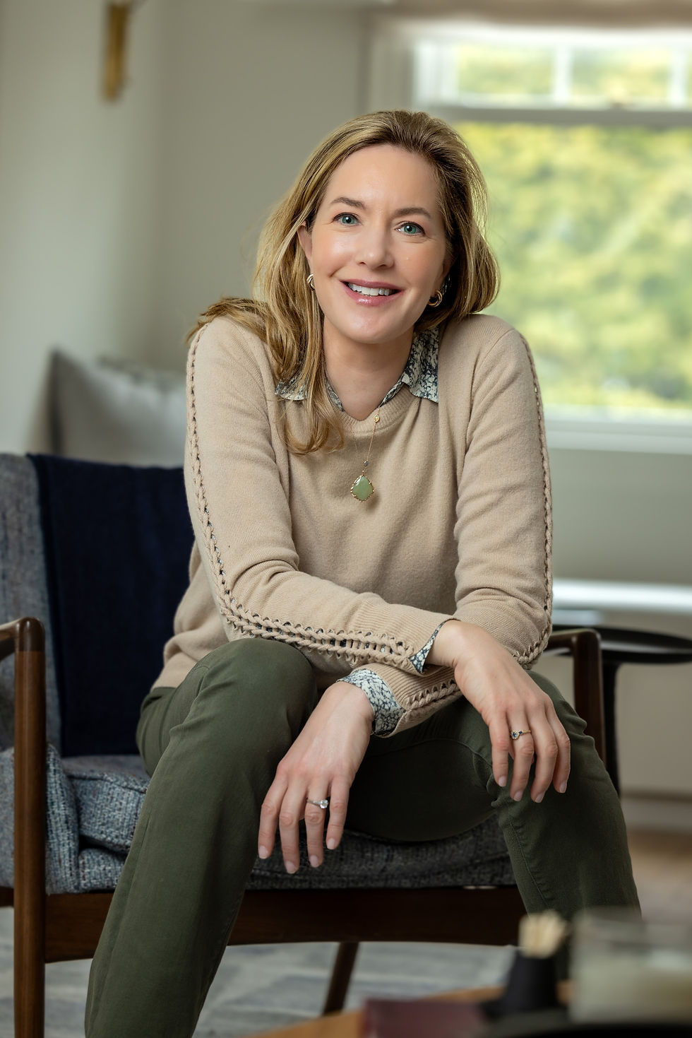 Woman in beige sweater and green pants smiles, seated in a cozy room with gray chair and window. Bright and relaxed atmosphere.