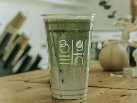 Plastic cup with green iced drink and white text logo on wooden table, blurred background with plant and objects. Calm, cozy atmosphere.