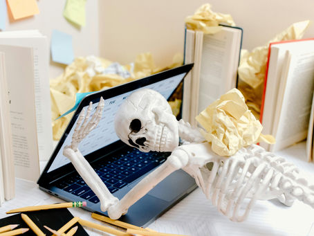 Skeleton sprawled over a laptop surrounded by open books, pencils, and crumpled papers. Sticky notes on the wall suggest chaos.