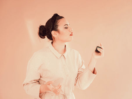 Woman in a cream outfit sprays perfume, eyes closed, against a light peach background, exuding a calm and elegant mood.