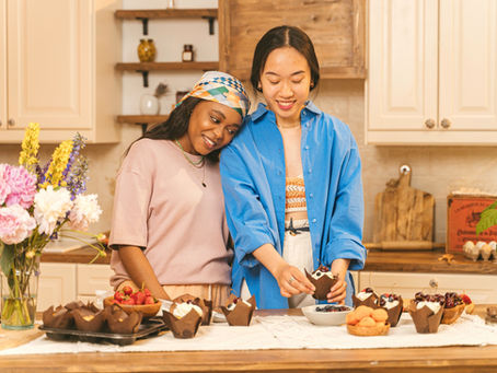 Two women in a kitchen, smiling while decorating cupcakes. A vase of flowers and bowls of fruit adorn the countertop. Bright and cozy setting.