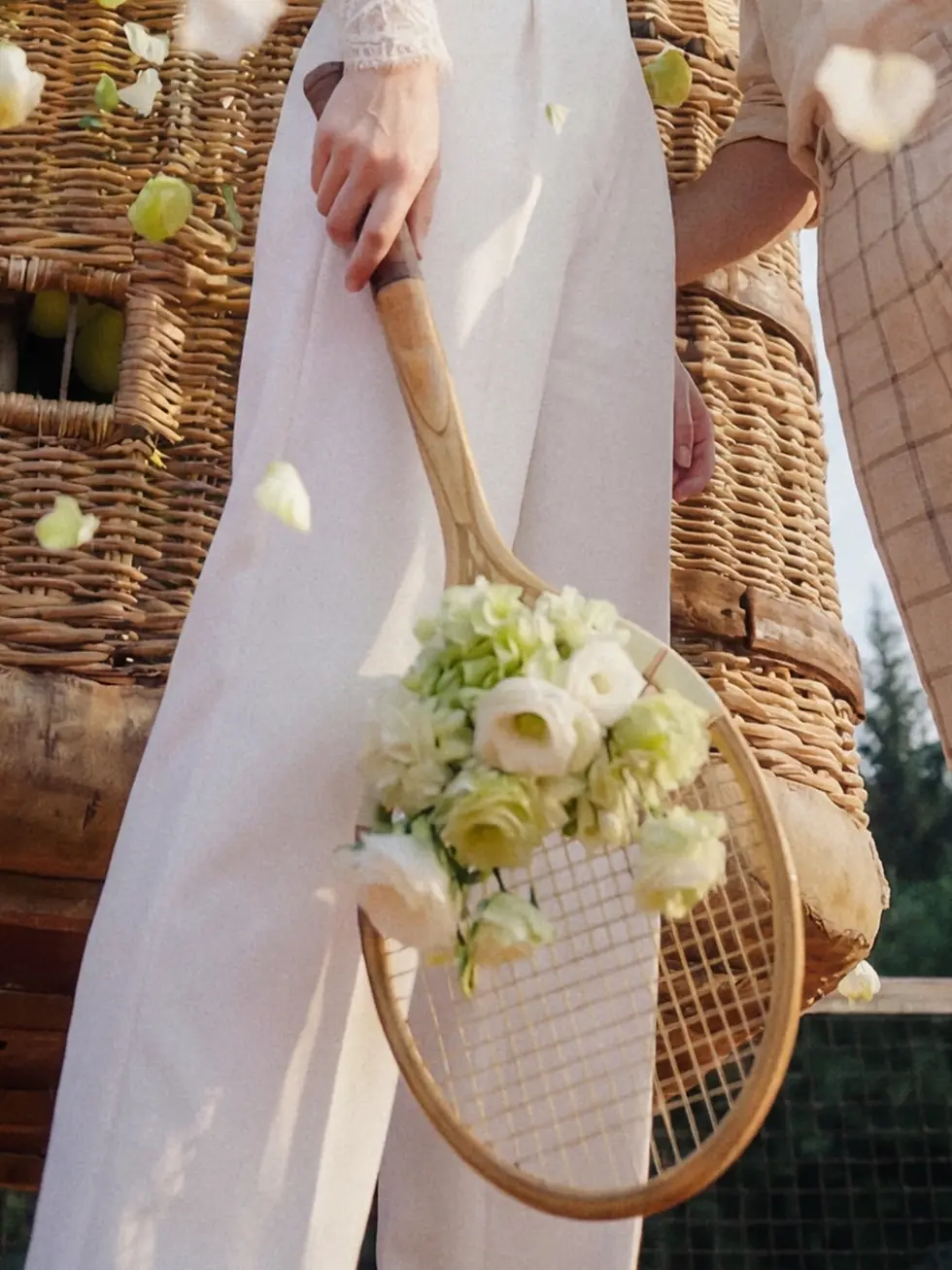 Detailshot auf einen vintage Tennisschläger und die Hose einer Braut in einem weißen Anzug. Im Hintergrund schwebt ein Heißluftballonkorb.