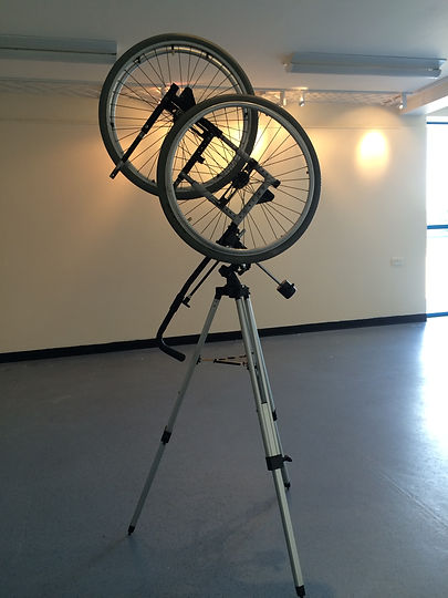 a side of a wheelchair and two wheels balanced on a tripod 6'3" in height in gallery setting crackedpainter. John Baldwin.