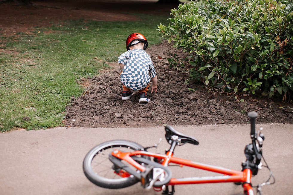 Child in checkered shirt, red helmet, squats on dirt by bushes. Orange bike lies on path. Green grass in the background. | family photos newcastle | Angela Fenwick Photography