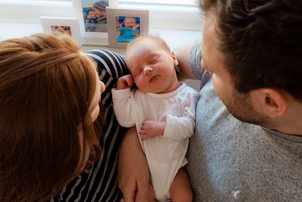 A couple lovingly holds a sleeping baby in a white outfit. Family photos in the background create a warm, intimate atmosphere. Baby Photoshoot Durham | Angela Fenwick Photography