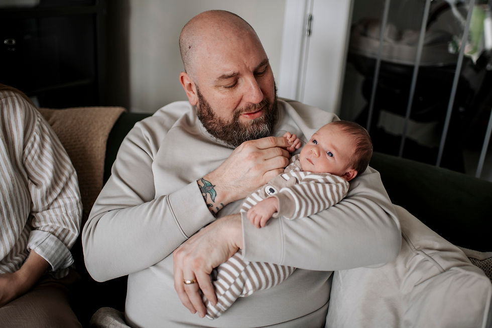 Bald man with a beard holds a baby in striped babygrow on a couch.