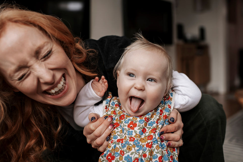 Woman and baby laugh indoors, baby sticks out tongue. Woman has red hair, smiling widely. Baby wears floral outfit. Natural family shoot at home Newcastle | Angela Fenwick Photography