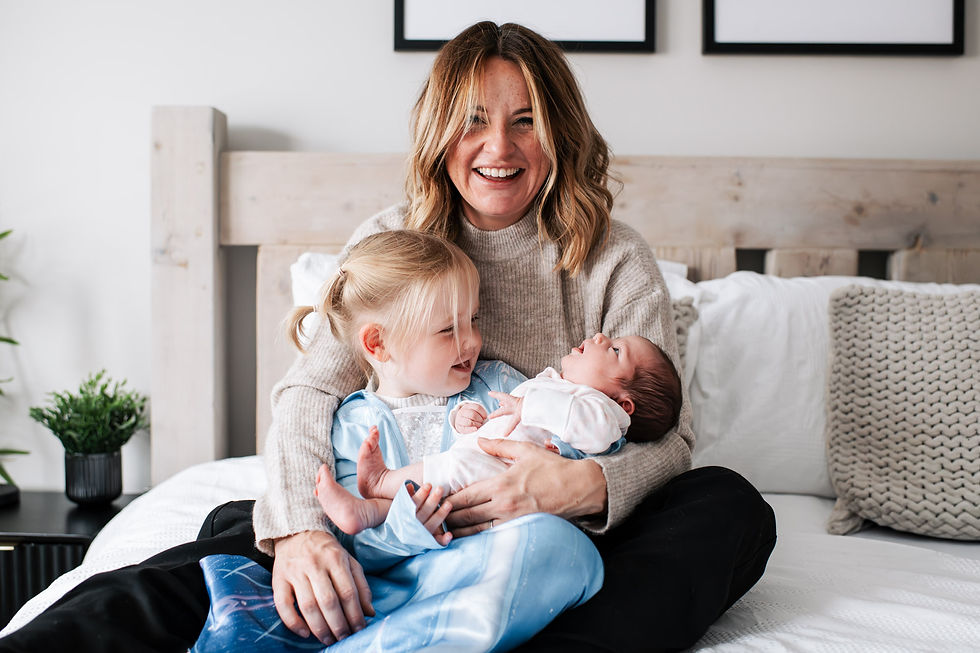 Woman holds smiling toddler and newborn on bed with neutral decor; cozy and joyful scene.Newborn shoot at home Durham. Angela Fenwick Photography