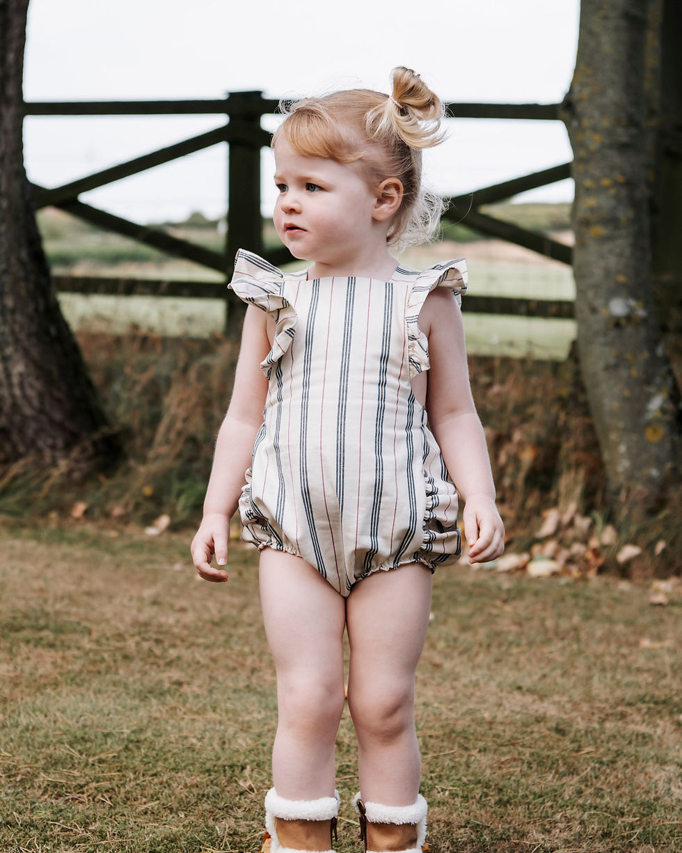 Toddler in a striped romper stands on grass, flanked by trees and a fence. She looks to the side, appearing curious. Earthy tones dominate.