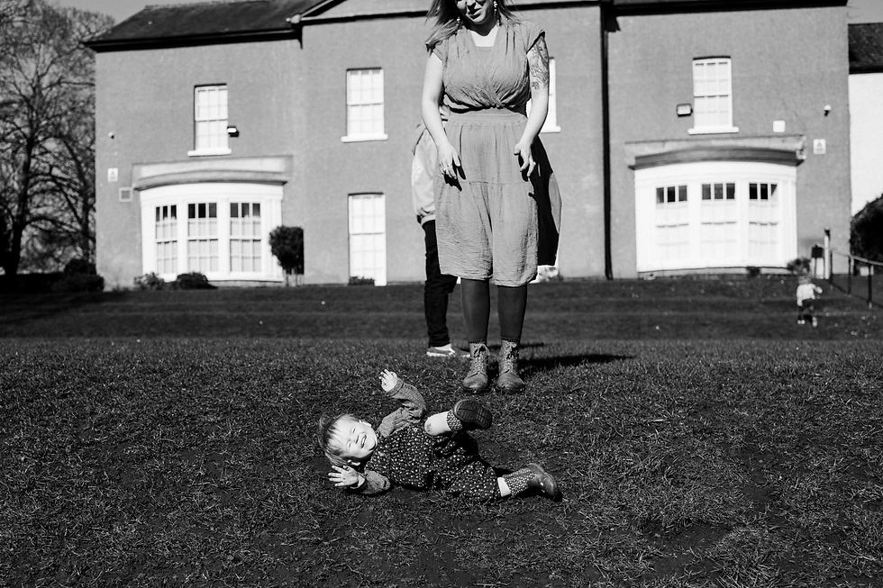 Toddler playfully lies on grass, woman stands nearby smiling. Background features a large house. Sunlit and relaxed atmosphere. Family documentary photography Durham and North East. Angela Fenwick Photography