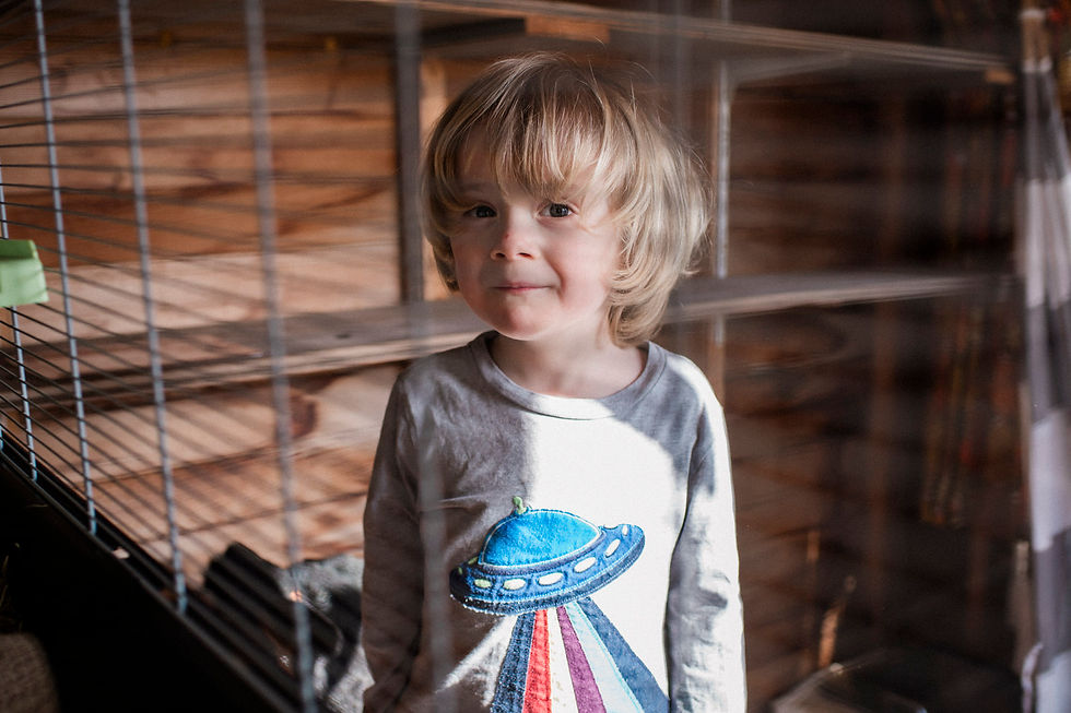 A child with blond hair, wearing a shirt with a colorful UFO print, stands indoors. A cage is visible in the foreground. Family documentary photography Durham and North East. Angela Fenwick Photography