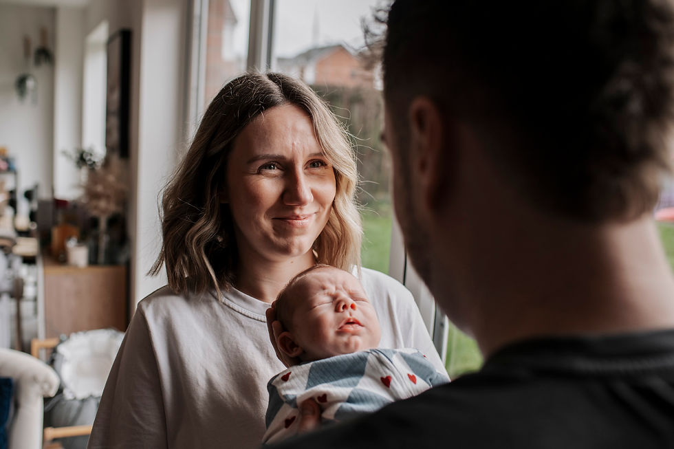 A woman smiles at a man holding a sleeping baby in a patterned blanket. They stand by a window in a cosy, softly lit room.Newborn photos at home Durham. Angela Fenwick Photography