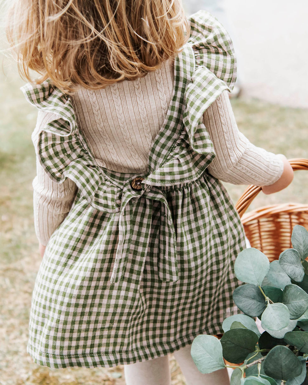 Young child holding a wicker basket, wearing a green checkered dress with ruffles in a grassy area. Cream sweater underneath, and eucalyptus leaves are visible.
