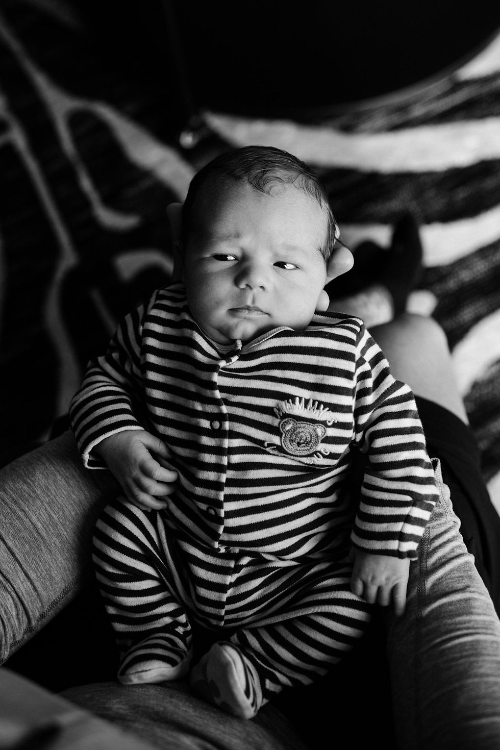 Baby in striped onesie lies on a person's lap. Black and white image with a patterned background. Natural newborn shoot at home Darlington | Angela Fenwick Photography