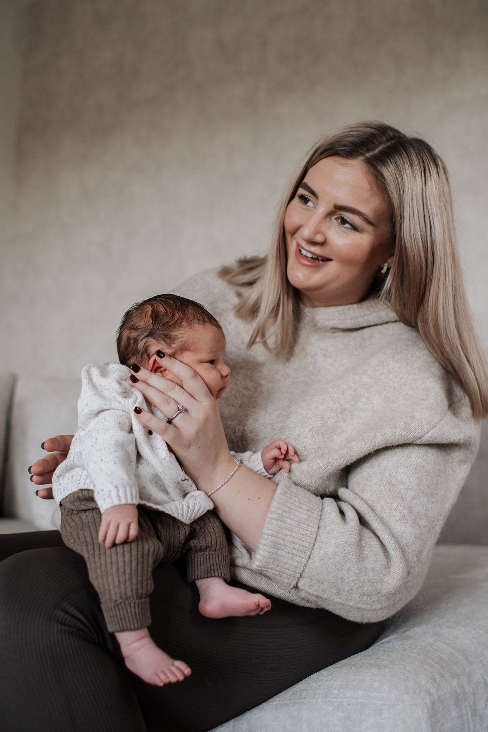 Woman holding a baby, smiling warmly. Both wear cozy knitwear in neutral tones. Soft, blurry background adds warmth to the scene.Natural newborn shoot at home Darlington | Angela Fenwick Photography